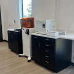 Dental lab equipment including a 3D printer and sterilizer on black cabinets in a modern workspace